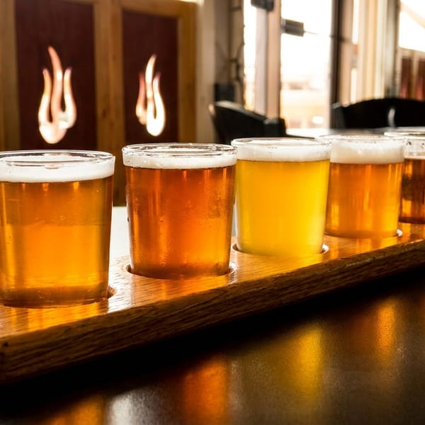 A wooden paddle holds five glasses of golden beer, with a restaurant interior in the background.