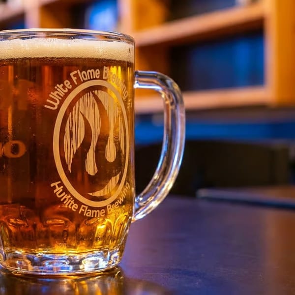 A frosty mug of White Flame Brewing beer with a foamy head sits on a dark table.