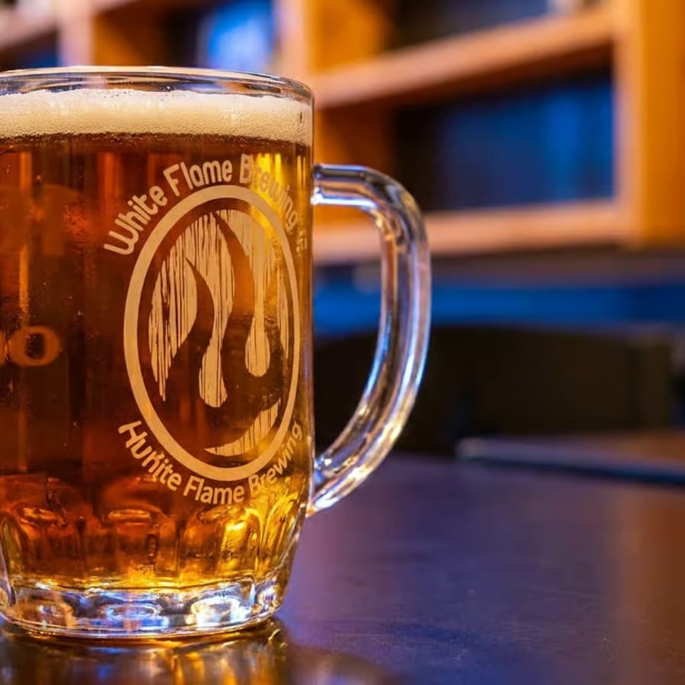 A frosty mug of White Flame Brewing beer with a foamy head sits on a dark table.