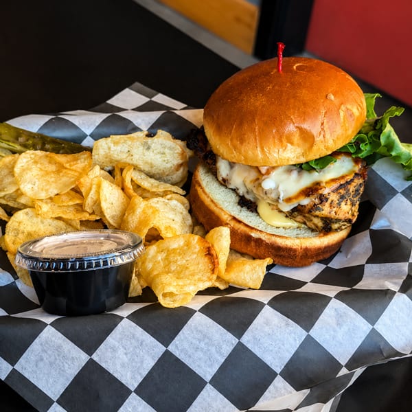 Grilled chicken sandwich with melted cheese, lettuce, potato chips, pickle spear, and dipping sauce.